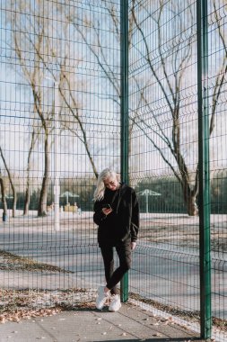 Açık havada akıllı telefon kullanırken basketbol sahasında poz veren mutlu ve çekici bir kadın.