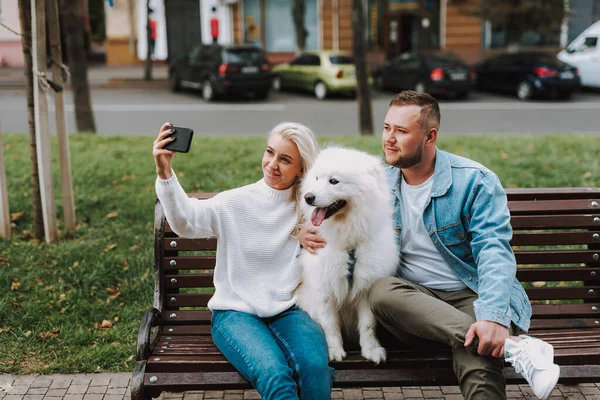 年轻的男女笑容满面地坐在一条小巷的长椅上 手里拿着一只白色毛绒绒的狗 用智能手机自拍 — 图库照片
