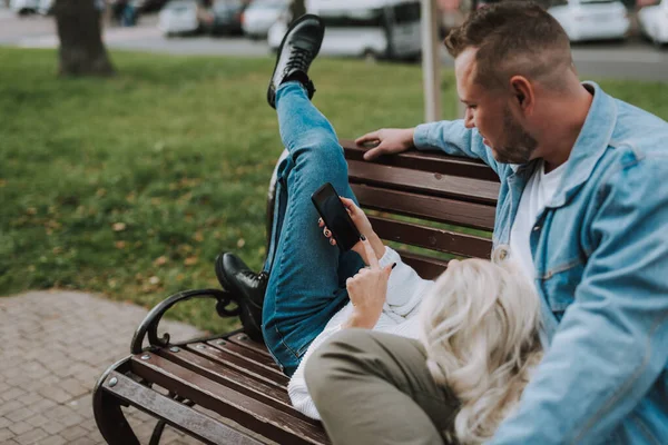 Młoda Blondynka Przegląda Zdjęcia Komórce Odpoczywa Swoim Facetem Ławce Alejce — Zdjęcie stockowe