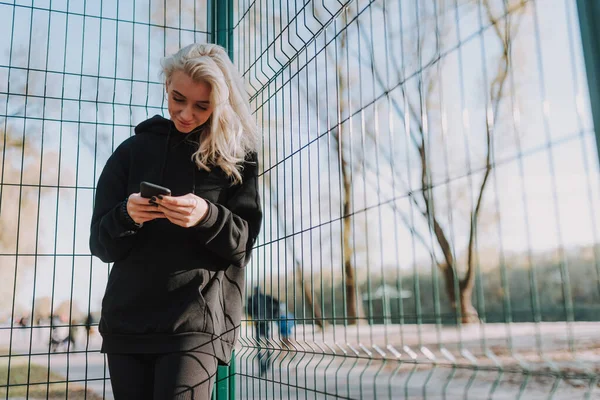 Waist up of happy pretty female wearing black sportswear standing on basketball court while using smartphone