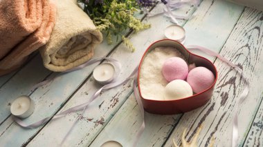 Kalp şeklinde bir kutuda, kokulu toplar ve banyo tuzu, mumların yanında, çiçekler ve havlular ahşap bir arka planda. Spa tedavileri için eşyalar.