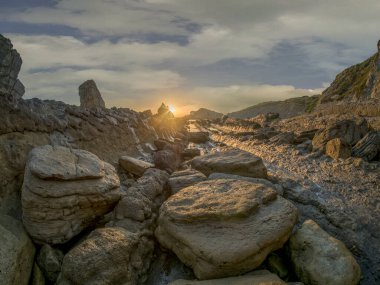 Sahilde gün doğumu, kayaların yanında güneş ve bulutların önünde.
