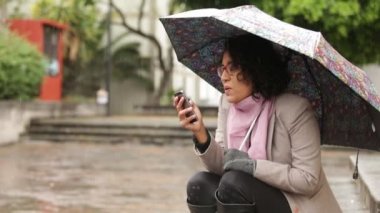 Esmer tenli, kıvırcık saçlı, yağmurda eğlenen, telefonlara cevap veren ve hayata gülümseyen Latin kız.