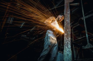 Fabrikadaki yakışıklı adam metali kesiyor, kıvılcımlar uçuşuyor, sahne ışıkları erkek spor fiziğine vuruyor.