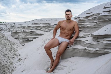 Beyaz şortlu seksi maço adam kumlu uçurumların kenarında oturuyor mavi gökyüzü arka planında, güneş tarafından aydınlatılmış fitness vücut geliştiricisi