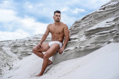 Beyaz şortlu seksi maço adam kumlu uçurumların kenarında oturuyor mavi gökyüzü arka planında, güneş tarafından aydınlatılmış fitness vücut geliştiricisi