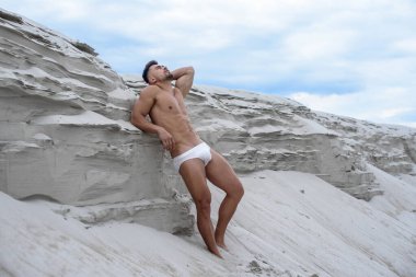 Seksi adam, beyaz maço adam, kumulların yanında duruyor. Mavi gökyüzü arka planına karşı seksi poz veriyor. Güneş tarafından aydınlatılan fitness vücut geliştiricisi.