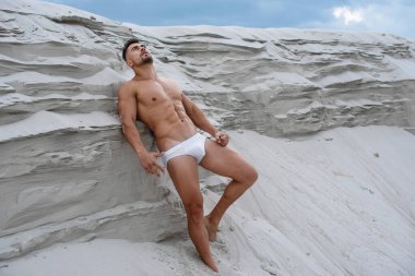Seksi adam, beyaz maço adam, kumulların yanında duruyor. Mavi gökyüzü arka planına karşı seksi poz veriyor. Güneş tarafından aydınlatılan fitness vücut geliştiricisi.
