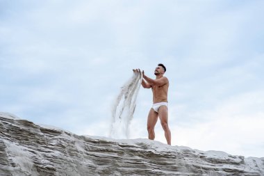 Beyaz şortlu seksi maço adam, kumların üzerinde mavi gökyüzüne karşı oturur. Arka planda kum tutar. Parmaklarının arasından kum tutar.