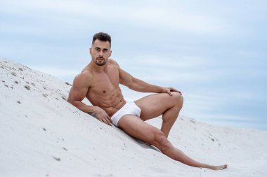 Seksi adam, beyaz maço adam, beyaz kumsalda uzanan gökyüzü arka planında, beyaz boş plaj, plajda güneşlenen sporcu.