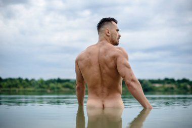 Seksi sporcu, maço vücut geliştirici, güzel bir suda sırtı kameraya dönük, arka planda bulutlu bir gökyüzü.