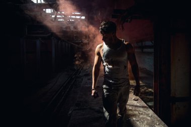 Katı kırmızı dumanlı bir adam iş yerinde yırtık bir tişörtle duruyor.