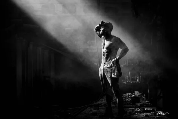 strong man against fog at factory exhausted stands, press and lateral abdominal muscles and pectoral muscles illuminated by light, place for text