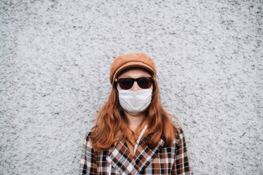 Koruyucu maskeli ve güneş gözlüklü bir kız duvarın yanında duruyor. 