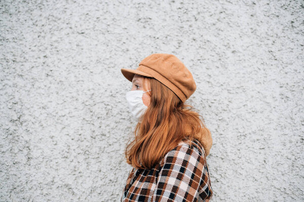 Redhead woman in a brown hat and a protective mask stands sideways against the background of the wall. isolated