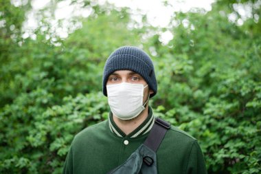 Koruyucu maskeli ve şapkalı bir adam yeşil bir çalılıkta duruyor. Kameraya bakıyorum.