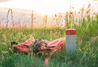 Kapalı termos ve kırmızı sırt çantası gökyüzünün altındaki çimenlerde yatıyor.