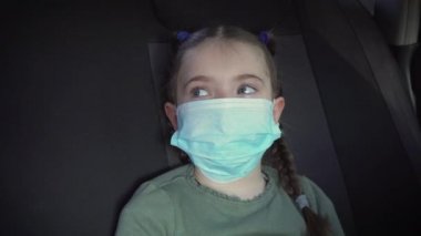 Coronavirus salgını ve kendini izole etme süresince şehir trafiğinde hareket halindeki arabanın arka koltuğunda koruyucu maskeli üzgün küçük kız.