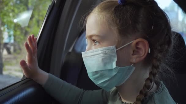 Retrato menina na máscara médica olha para fora janela do carro em movimento na rua. Auto-isolamento em quarentena — Vídeo de Stock