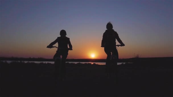 Siluetas de dos miembros de la familia madre e hijo montando sus bicicletas de montaña cerca del lago en increíbles rayos de puesta de sol en cámara lenta — Vídeo de stock