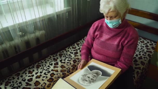 Senior mujer caucásica en máscara protectora en su cara mirando su retrato en la foto en su habitación en casa — Vídeo de stock