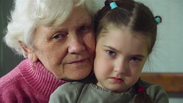 Portrait of old woman granny with her sad granddaughter hug and looks at camera at room. Warm relations between two generations — Stock Video