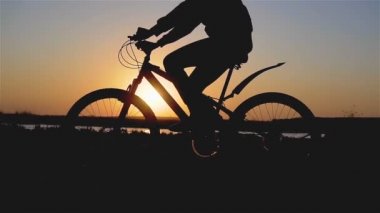Genç bir kadın ya da kızın nehir sahilinde bisiklet sürerken ve gün batımında pedal çevirirken güzel bir bahar fotoğrafı.