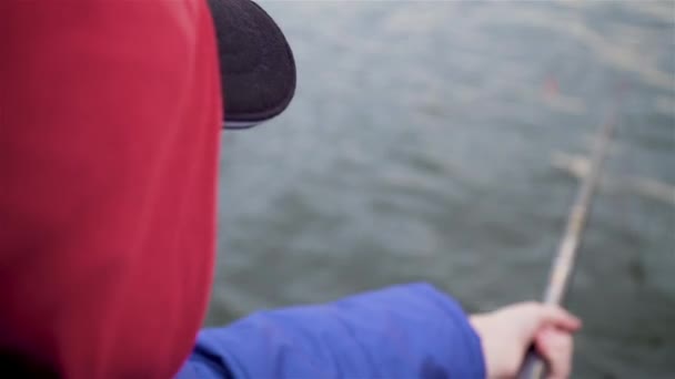Κοντινό πλάνο του αγοριού που ψαρεύει στο ποτάμι με καπέλο και κουκούλα και μπλε σακάκι — Αρχείο Βίντεο