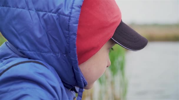 Jonge jongen in kap en kap kijkt naar vishengel drijven op de rivier in de koude herfst bewolkt weer — Stockvideo