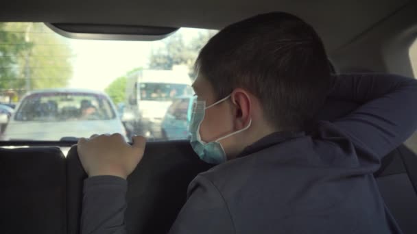 Ein kleiner Junge in medizinischer Schutzmaske blickt aus dem Autofenster auf den Stadtverkehr. Selbstisolierung in Coronavirus-Quarantäne — Stockvideo