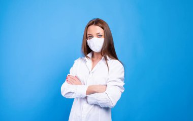 Tek kullanımlık yüz maskesi olan beyaz tişörtlü güzel beyaz kadın. Virüs ve enfeksiyona karşı koruma. Stüdyo portresi, mavi arka planlı konsept. Kollar göğüsten bağlanmış..