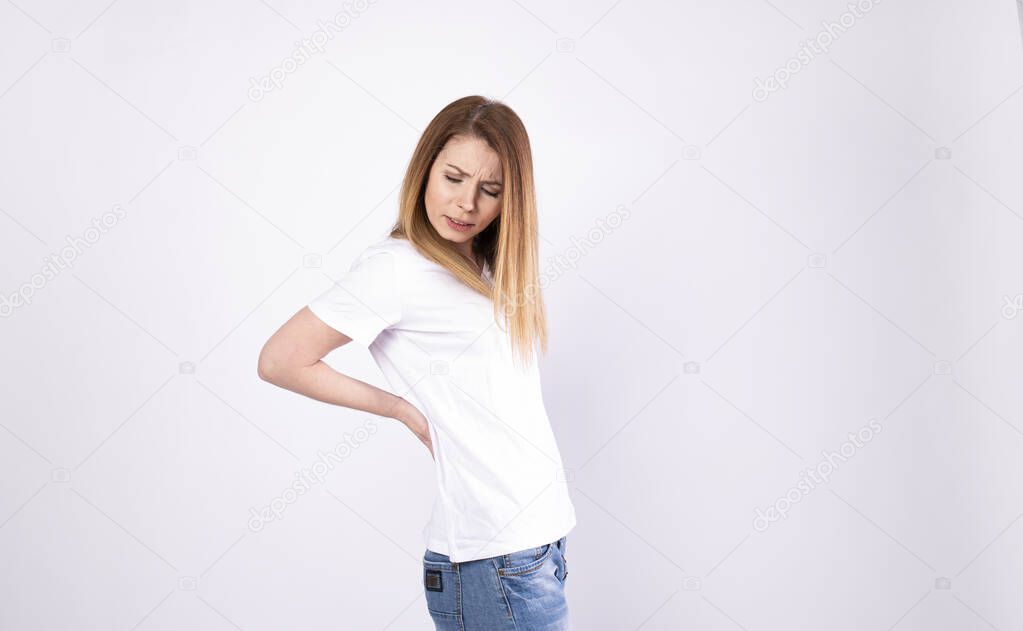 Young woman in a white pullover with back ache clasping her hands to her lower back as she looks up wincing in pain, side view.