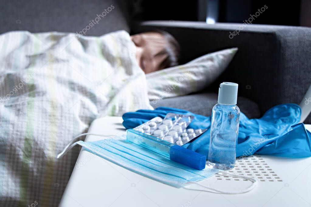 Sick child covered with a blanket is lying in bed. Next to tablets, protective mask, thermometer, gloves and bottle hand sanitizer on the chair