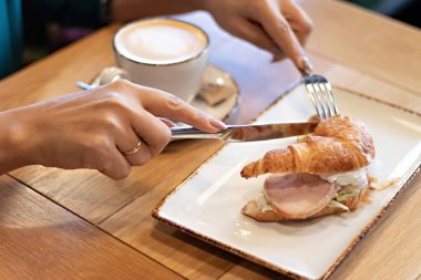 Kız kahvaltı yapıyor ve bir restoranda sandviç kesiyor, yakın çekim. Kahve ve kruvasan içen kadın.