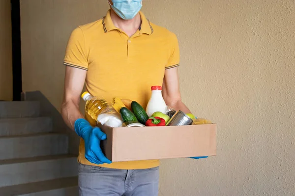Man in protective mask and gloves holds box of food. Donation box with food supply, canned food, cereals, vegetables. Concept of food delivery