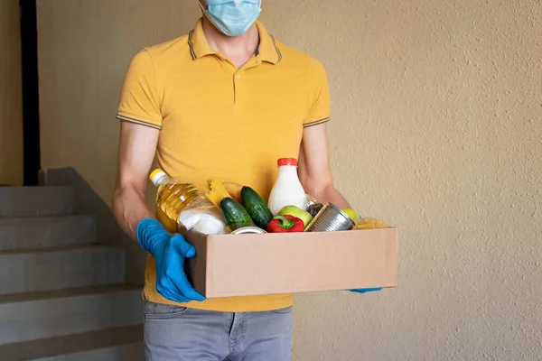 Koruyucu maskeli ve eldivenli adam yiyecek dolu bir kutu tutuyor. Bağış kutusunda yiyecek, konserve yiyecek, tahıl, sebze. Yemek teslimatı kavramı