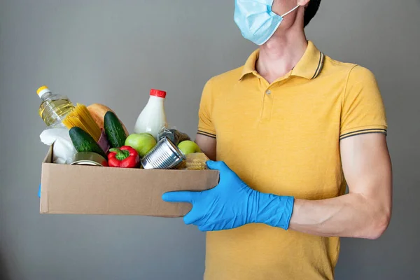 Koruyucu maskeli ve eldivenli adam bir kutu yiyecek tutuyor. Bağış kutusunda yiyecek, konserve yiyecek, tahıl, sebze. Yemek teslimatı kavramı