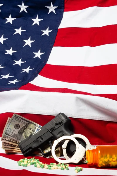 Gun, pills, handcuffs and cash in front of the American flag, vertical