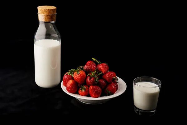 Bir şişe süt, bir tabak çilek, siyah arka planda bir bardak süt..