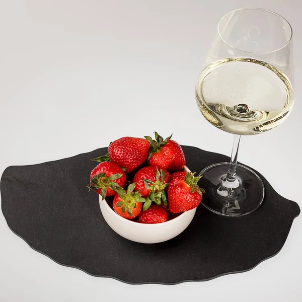 Beyaz bardakta taze çilek, beyaz arka planda siyah taş tepside bir bardak beyaz şarap.
