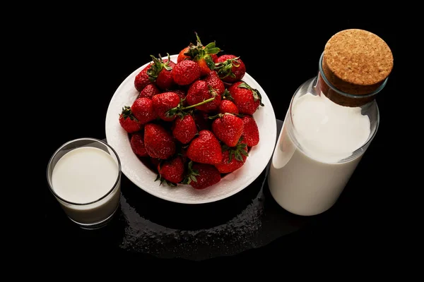 Placa blanca con fresas, una botella de leche y un vaso de leche sobre fondo negro — Foto de Stock