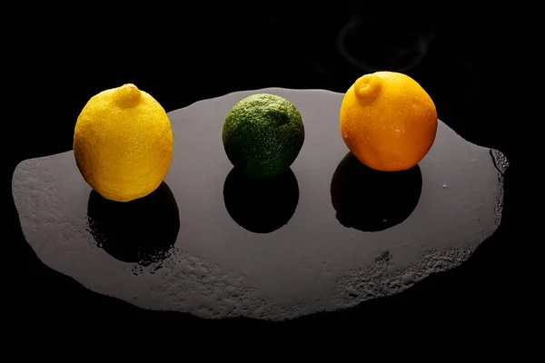 Dwie żółte cytryny i zielona limonka na mokrej kamiennej tacy na czarnym tle. — Zdjęcie stockowe