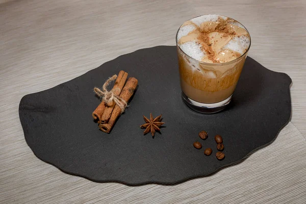 Tarçın çubukları ve siyah taş tepside bir kova olan cam bardakta Dalgona kahvesi.