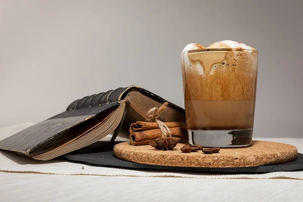Caffè Dalgon in una tazza di vetro su uno stand di sughero accanto a un libro, cannella, chicchi di caffè e badyan su un vassoio di pietra nera su uno sfondo leggero . — Foto Stock