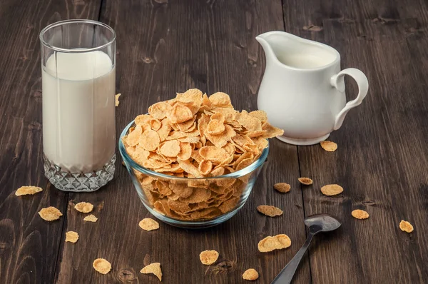 Corn flakes with milk jug and glass on wooden table — Stock Photo, Image