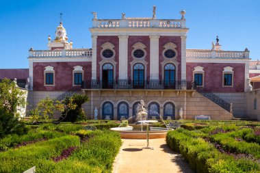 Duvarların açık bordo rengi, beyaz detaylar. Estoi Sarayı çeşmeli bir bahçeyle çevrili. Parlak mavi gökyüzü. Estoi, Portekiz.