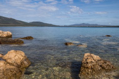 Akdeniz 'in temiz, temiz ve sakin suları, kireçtaşı kayalarıyla. Ufuktaki dağlar. Baharda açık beyaz bulutlu mavi gökyüzü. Sardunya 'nın batı kıyısı, İtalya.
