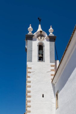 Igreja Matriz 'in çan kulesi ve saati olan beyaz cephesi. Horoz tepede. Parlak mavi gökyüzü. Estoi, Portekiz.