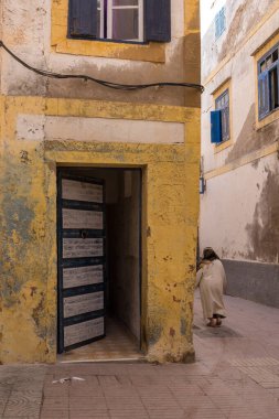 Yıpranmış sarı cepheli ve kapısı açık bir evin köşesi. Geleneksel giyinmiş bir kadın sokağa çıkıyor. Essaouira, Fas 'ın eski kenti (Medina).