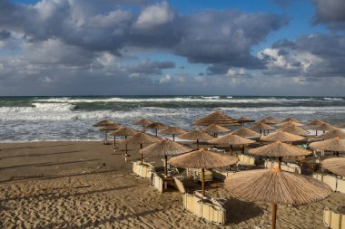 Sonbaharda yağmurlu ve fırtınalı bir günde, kumsalda bir grup şezlonglu ve güneşli şemsiyeli bir kumsal. Deniz dalgaları ve yoğun renkli bulutlar. Retimnon, Girit, Yunanistan.